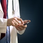 Male doctor in white coat is using a modern smartphone device with tocuh screen.